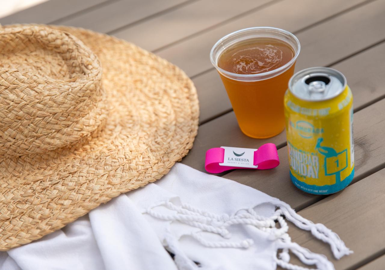 A craft beer can, a beach hat and a La Siesta wristband set up by a beach towel.
