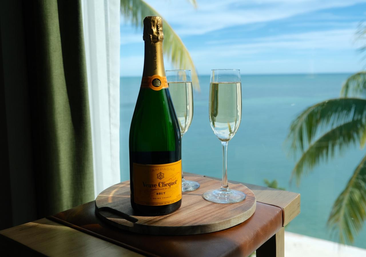 A wooden table with a bottle of champagne and two glasses. The table is set in front of a window overlooking the ocean. 