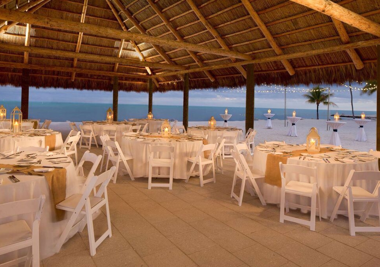 large tiki on the beach set up for a gathering with white tables and chairs